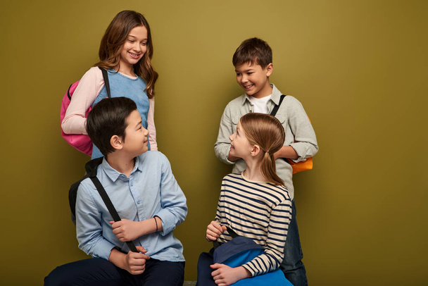 Позитивные мультиэтнические школьники разговаривают с друзьями-подростками, держа рюкзаки, глядя друг на друга и отмечая международный день детей на фоне хаки - Фото, изображение