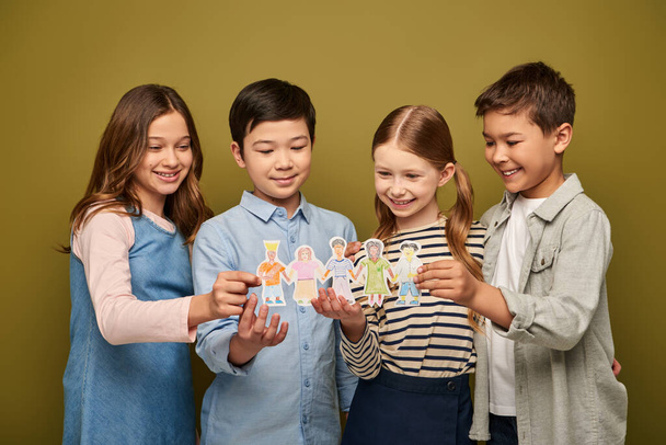 Smiling interracial preteen kids in casual clothes with friends holding drawn paper characters while celebrating child protection day on khaki background - Foto, imagen