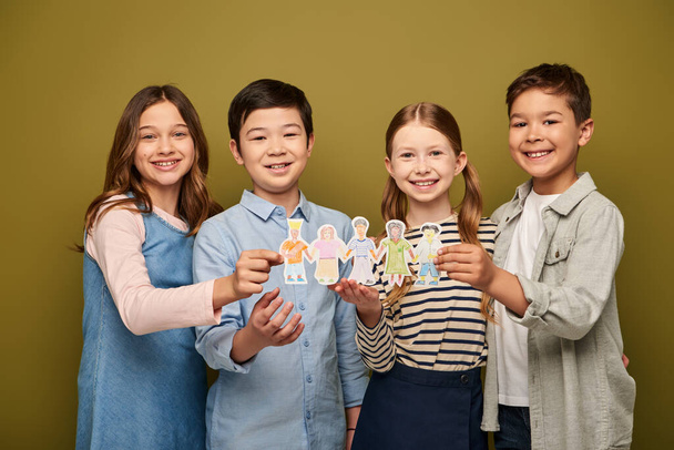 Детишки в повседневной одежде, держащие бумажных персонажей и улыбающиеся перед камерой во время празднования Международного дня защиты детей на фоне хаки - Фото, изображение