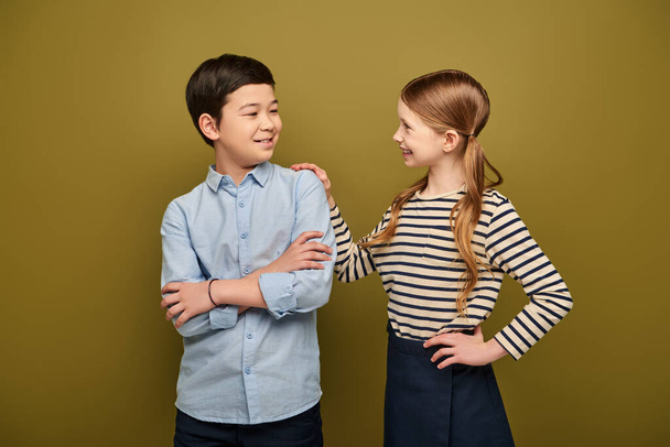 Smiling redhead preteen girl hugging and looking at asian friend folding hands during international child protection day celebration on khaki background - Фото, зображення