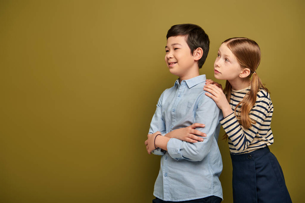 Redhead girl in striped blouse hugging smiling asian friend crossing and and looking away during child protection day celebration on khaki background with copy space  - Φωτογραφία, εικόνα