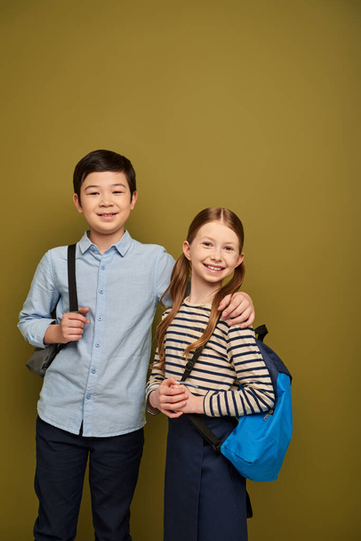 Smiling asian boy with backpack hugging red haired friend and looking at camera during international child protection day celebration on khaki background - Φωτογραφία, εικόνα