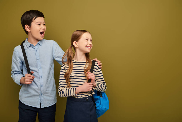 Excited asian boy with opened mouth with backpack opening mouth and hugging cheerful redhead friend and looking away during child protection day celebration on khaki background - Photo, Image