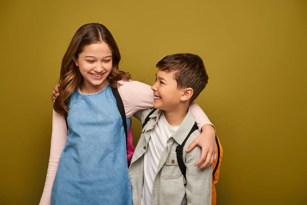 Carefree preteen and multiethnic kids in casual clothes with backpacks hugging each other and smiling during global child protection day on khaki background - Photo, Image