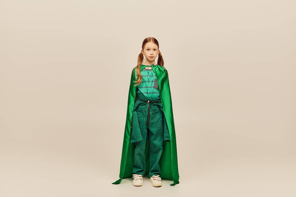 Rousse fille préadolescente en costume de super-héros vert et cape regardant la caméra tout en se tenant sur fond gris pendant la célébration de la journée mondiale de la protection de l'enfance  - Photo, image