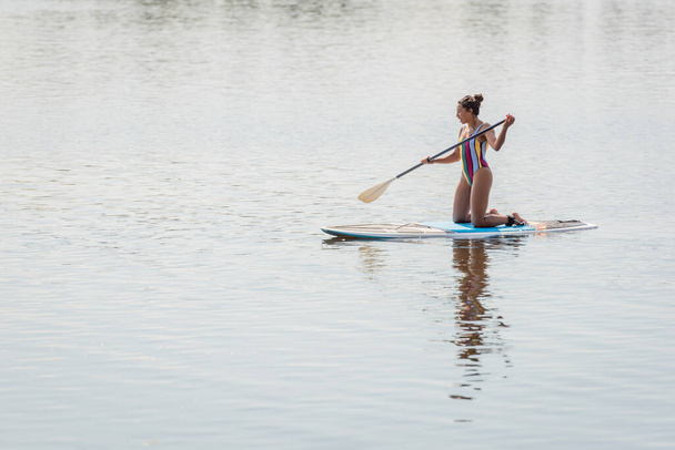 schlanke afrikanisch-amerikanische Frau im bunten Badeanzug mit Paddel, während sie auf Knien steht und am Wochenende an einem Sommertag auf einem Sup Board auf dem Fluss segelt - Foto, Bild