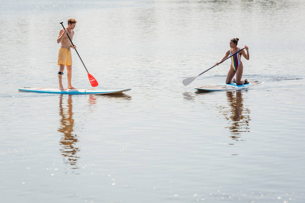 sportive african american woman in colorful swimsuit and young redhead man in swim shorts spending summer weekend while sailing on sup boards together - Fotó, kép