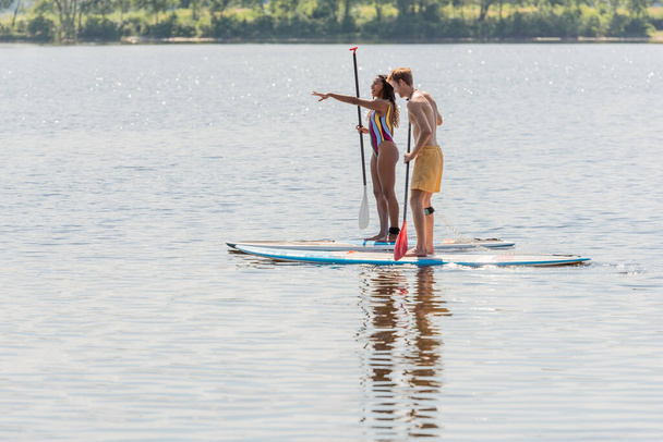 vista lateral de la mujer afroamericana en traje de baño colorido apuntando con la mano cerca de hombre joven deportivo mientras navega en tablas de sup durante el fin de semana en el lago - Foto, Imagen