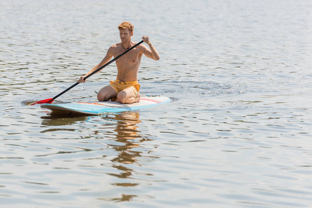 young and sportive redhead man in yellow swim shorts spending summer weekend on river while sitting on sup board and sailing with paddle during water recreation - Photo, Image