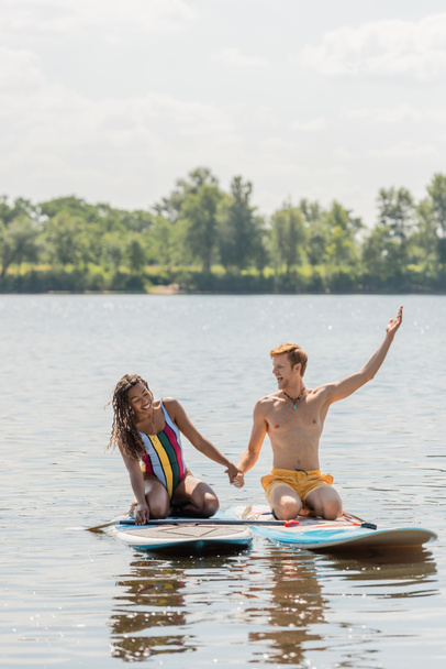 carefree redhead man holding hands with cheerful african american woman and pointing away while sitting on sup boards on lake with green picturesque bank - Photo, image