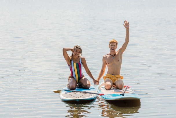 насолоджений рудий чоловік тримає руки з усміхненою афроамериканкою в смугастому купальнику і показує жест привітання з дощок на річці влітку
 - Фото, зображення