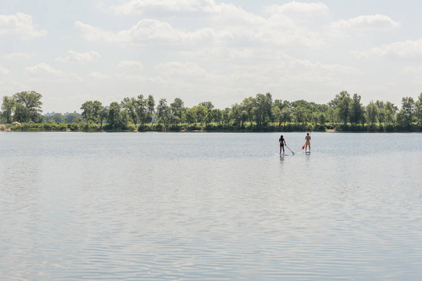 widok z daleka od międzyrasowych i aktywnych para spędzając letni weekend podczas żeglowania na deskach supsup z wiosłami na jeziorze z malowniczym brzegu pod chmurnym niebem - Zdjęcie, obraz