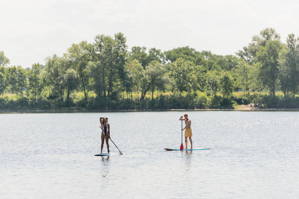 full length of sportive african american woman with young friend sailing on sup boards with paddles along riverside with green trees during summer weekend - Foto, imagen