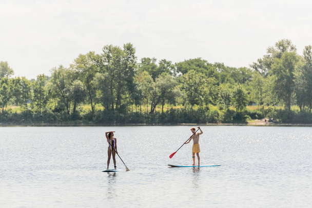 full length of african american woman sailing on sup board and looking away near young and sportive man in swim shorts on scenic lake with green trees on shore - Photo, Image