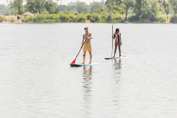 full length of young sportive man and african american woman in striped swimsuit spending time on lake by sailing on sup boards with paddles on summer weekend - Photo, image