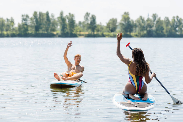 brunette african american woman in striped swimsuit and overjoyed redhead man waving hands at each other while sailing on sup boards on lake in summer - Photo, Image