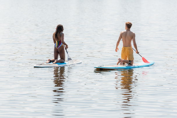 задний вид спортивного и рыжеволосого мужчины и африканской американки в полосатых купальниках, стоящих на коленях на досках и плывущих по реке в день летних каникул  - Фото, изображение