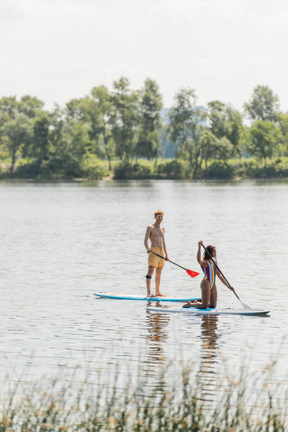 redhead and sportive man paddleboarding along green riverside near african american woman in colorful swimsuit during summer recreation on summer weekend - Foto, Bild