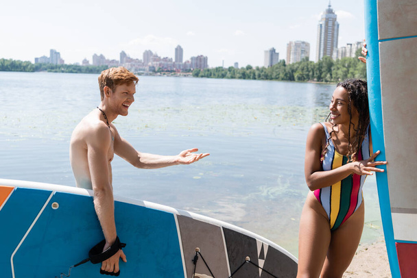 happy redhead man holding sup board and talking to enchanting african american woman in colorful swimsuit near lake and cityscape on background in summer - Photo, Image