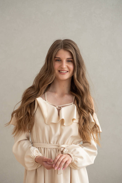 Hermosa joven mujer sonriente retrato con el pelo ondulado, vestido con un vestido beige posando en el interior  - Foto, imagen