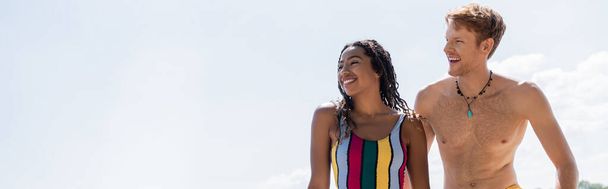 przepełniona radością afrykańska kobieta w kolorowym stroju kąpielowym i młody rudy mężczyzna odwracając wzrok i uśmiechając się pod błękitnym niebem w letni weekend na świeżym powietrzu, sztandar - Zdjęcie, obraz