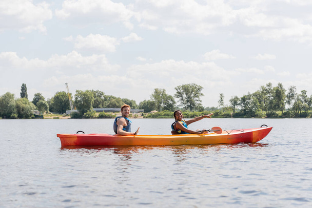 carefree african american woman in life vest pointing ahead and smiling at camera while sailing in sportive kayak with redhead friend during summer weekend on river - Photo, Image