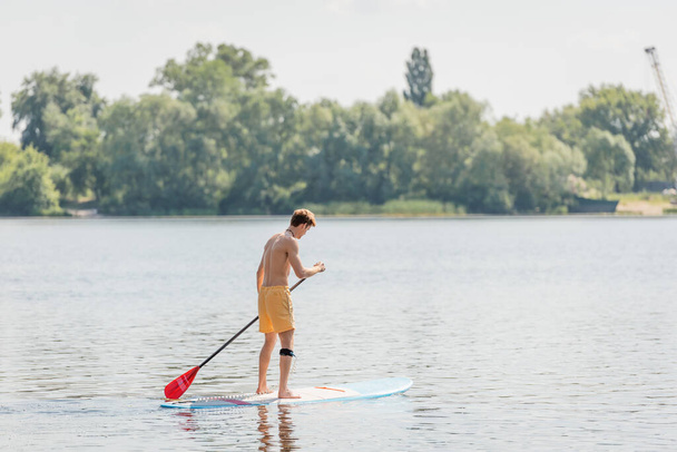 volle Länge des rothaarigen Mannes in gelben Badehosen, der auf einem Surfbrett mit Paddel segelt, während er am Sommerwochenende Zeit auf einem Fluss mit grünem malerischem Ufer verbringt - Foto, Bild