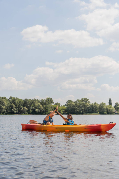 despreocupados pareja interracial en chalecos salvavidas dando cinco altos durante el fin de semana de verano en el río y navegar en kayak deportivo a lo largo de la orilla verde del río bajo el cielo azul con nubes blancas - Foto, Imagen