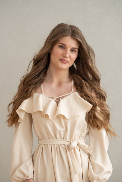 Beautiful young smiling woman portrait with wavy hair, dressed in a beige dress posing in the interior  - Foto, immagini