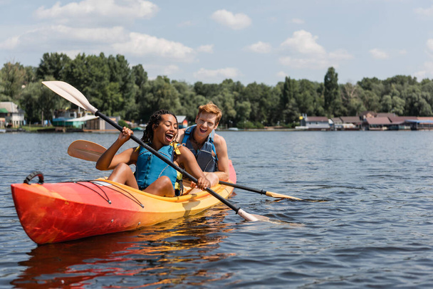 mujer afroamericana llena de alegría y joven pelirrojo en chalecos salvavidas pasar el fin de semana de verano navegando en kayak con remos en el lago con paisaje verde orilla - Foto, imagen