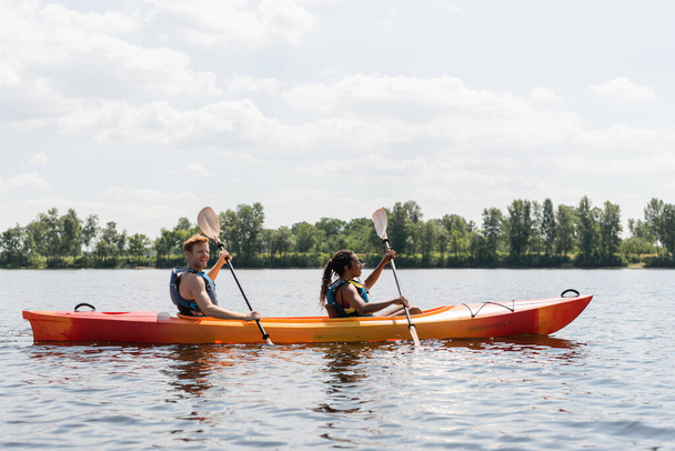 vista lateral de sonriente pelirroja hombre y morena afroamericana mujer en chalecos salvavidas pasar el fin de semana de verano navegando en kayak en el río bajo el cielo nublado en verano - Foto, imagen
