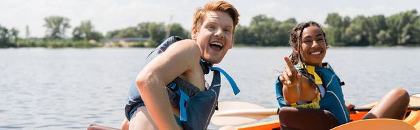 giovane ed eccitato uomo rossa con la bocca aperta guardando la fotocamera vicino gioia donna afroamericana in giubbotto di salvataggio che punta con il dito durante la navigazione in kayak il giorno d'estate, banner - Foto, immagini