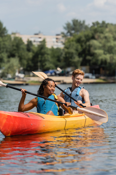 pareja interracial sin preocupaciones en chalecos salvavidas sosteniendo paletas y mirando el agua clara mientras navega en kayak deportivo durante el fin de semana de recreación en el día de verano - Foto, Imagen