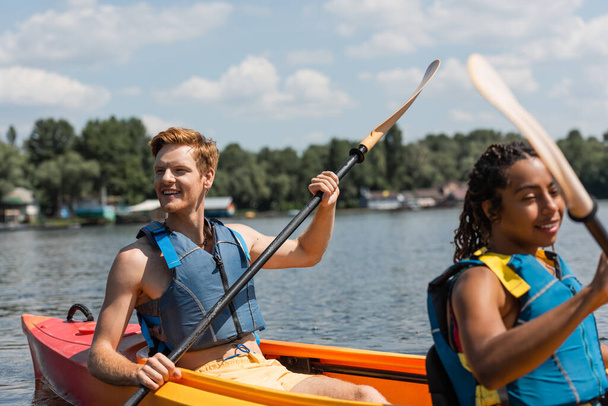 hombre pelirrojo excitado en chaleco salvavidas mirando hacia otro lado mientras navega en kayak deportivo con bastante mujer afroamericana durante el ocio recreativo en el lago en verano - Foto, Imagen