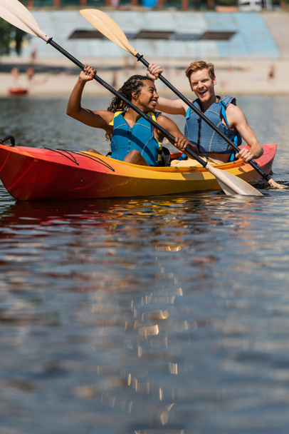 encantadora mujer afroamericana en chaleco salvavidas mirando al hombre pelirrojo excitado mientras navega en kayak deportivo con paletas en el día de verano en primer plano borrosa - Foto, Imagen