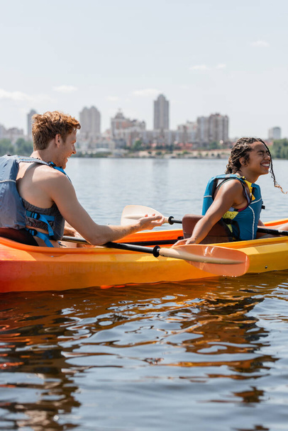 giovane uomo rossa in giubbotto di salvataggio parlando con la donna afro-americana felicissima ridendo ad occhi chiusi e seduto in kayak sportivo sul fiume con paesaggio urbano offuscato sullo sfondo - Foto, immagini
