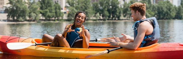 mulher americana africana alegre em colete de vida gesticulando perto jovem ruiva homem enquanto passar o fim de semana de verão no rio e sentado em caiaque esportivo, bandeira - Foto, Imagem