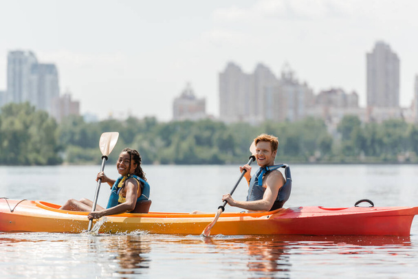 enchanting african american woman and sportive redhead man in life vests looking at camera during sailing in kayak along riverside with blurred cityscape - Photo, Image