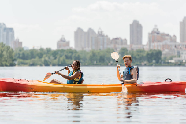 positive and sportive interracial couple in life vests sitting with paddles in kayak and smiling at camera on river with picturesque cityscape on blurred background - Photo, Image