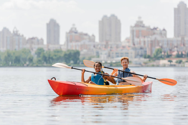 amigos multiétnicos felices e impresionados en chalecos salvavidas sosteniendo paletas y sonriendo a la cámara en kayak deportivo en el río con paisaje urbano escénico sobre fondo borroso - Foto, Imagen