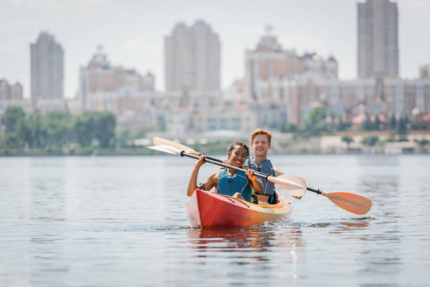spensierata coppia interrazziale in giubbotti di salvataggio in possesso di pagaie e guardando la fotocamera durante la navigazione in kayak sportivo sul lago con paesaggio urbano offuscato sullo sfondo - Foto, immagini