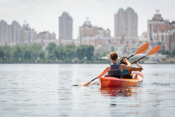 arrière vue du couple multiethnique actif dans les gilets de sauvetage pagayant en kayak sportif sur la surface de l'eau calme le jour d'été avec vue sur les bâtiments urbains flous sur le bord de la rivière  - Photo, image