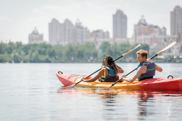 redhead man and african american woman in life vests spending time on river by sailing in sportive kayak and enjoying picturesque cityscape on blurred background - Photo, Image