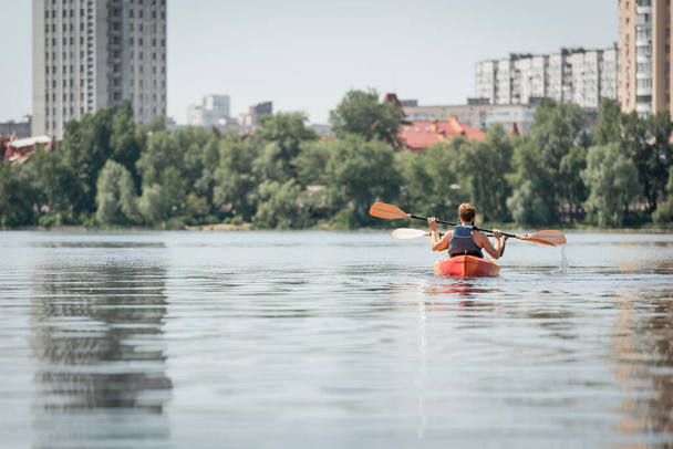 vista trasera de la pareja interracial deportiva en chalecos salvavidas que navegan en kayak cerca de la orilla del río con árboles verdes y edificios modernos de la ciudad durante el fin de semana de verano - Foto, imagen