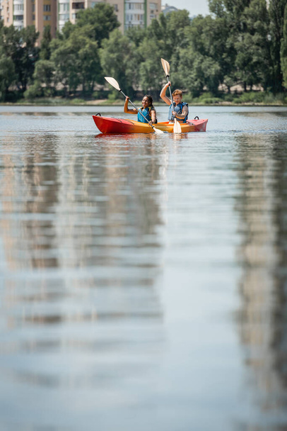 young active man and african american woman in life vests sailing in sportive kayak with paddles on blurred water surface with green riverside on background - Photo, Image