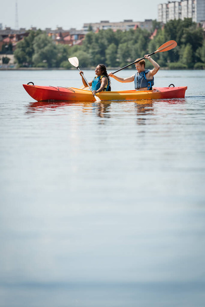 happy african american woman and young active man in life vests spending time on city lake and sailing in sportive kayak with paddles on blurred foreground - Photo, Image
