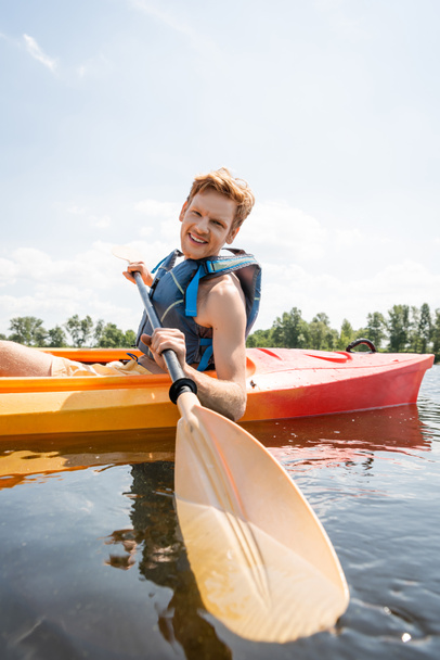 cheerful and redhead man in life vest holding paddle and looking at camera while spending time on lake by sailing in kayak during water recreation on summer weekend - Photo, Image