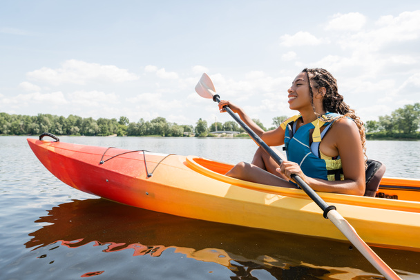 vista laterale di spensierata e attiva donna afroamericana in giubbotto salvagente tenuta pagaia durante la navigazione in kayak sul lago con verde pittoresco riva in estate - Foto, immagini