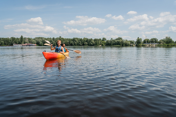active multiethnic couple in safe vests sailing in sportive kayak on picturesque lake with green shore under blue and cloudy sky during water recreation on summer weekend - Photo, Image