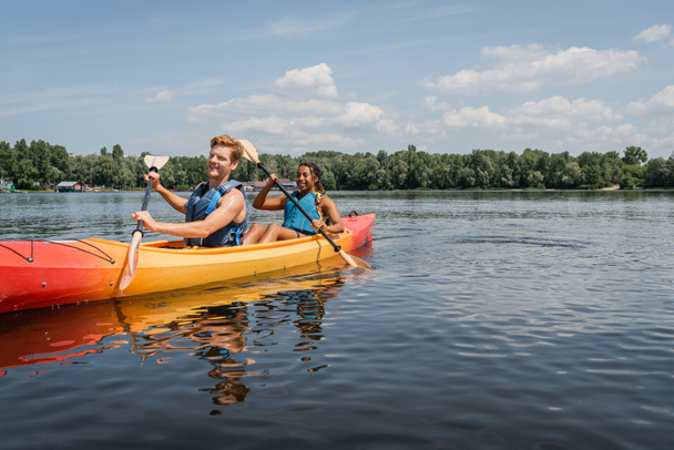 glückliches und sportliches gemischtes Paar in Schwimmwesten, das seinen Urlaub am malerischen See verbringt und im Kajak mit Paddeln unter blauem Himmel mit Wolken segelt - Foto, Bild